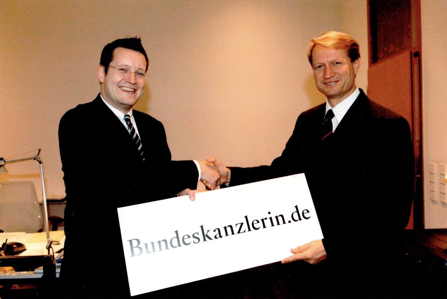Lars M. Heitmüller und Ulrich Wilhelm geben sich die Hand und halten gemeinsam ein Schild mit der Aufschrift Bundeskanzlerin.de